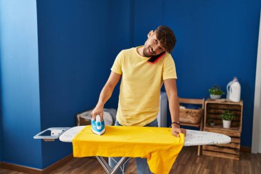 Young arab man talking on smartphone ironing clothes at laundry room