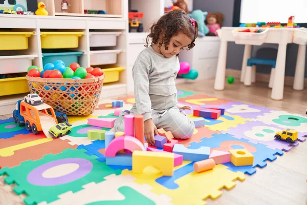Adorable hispanic girl playing with construction blocks sitting on floor at kindergarten