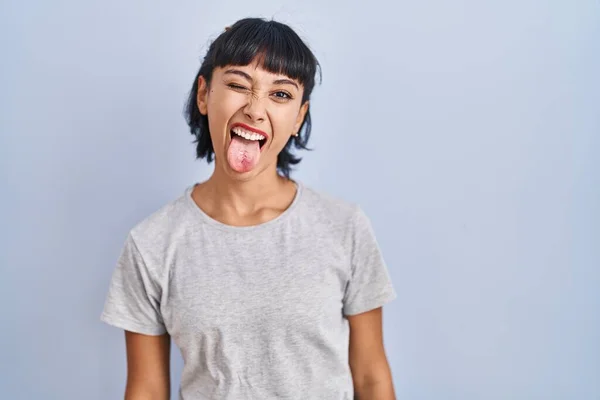 青い背景にカジュアルなTシャツを着た若いヒスパニック系の女性が面白い表情で幸せ舌を突き出しています 感情の概念 — ストック写真