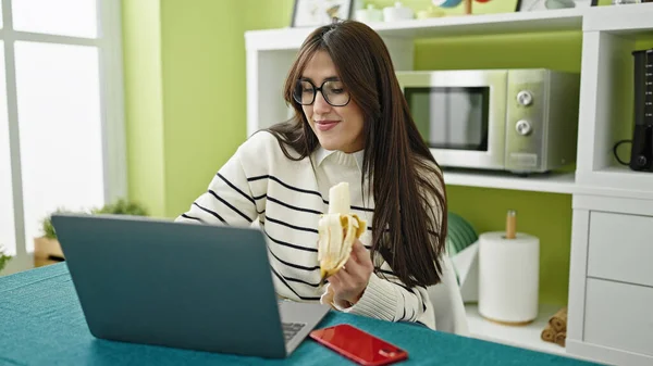 年轻美丽的惊慌失措的女人在饭厅用笔记本电脑吃香蕉 — 图库照片