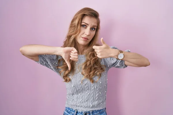 Beautiful blonde woman standing over pink background doing thumbs up and down, disagreement and agreement expression. crazy conflict