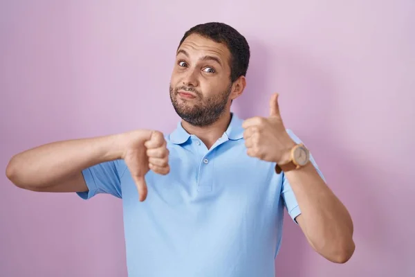 Hispanic man standing over pink background doing thumbs up and down, disagreement and agreement expression. crazy conflict
