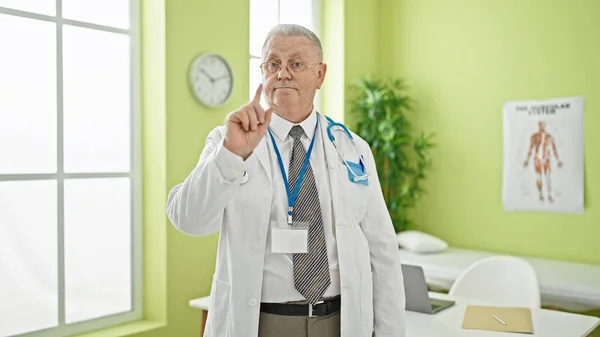 Middle age grey-haired man doctor standing with serious expression saying no with finger at clinic
