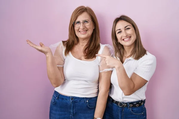 ヒスパニック系の母親と娘を身に着けていますカジュアルな白いTシャツ上ピンクの背景驚きと笑顔にカメラを提示しながら手と指差し指 — ストック写真
