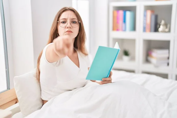 若いですCaucasian女性座っていますベッドの上に自宅で本を読んで指でカメラを指して あなたに 自信を持ってジェスチャーを真剣に見て — ストック写真
