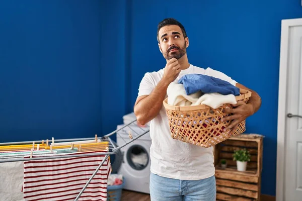 Young Hispanic Man Beard Holding Laundry Basket Serious Face Thinking — Stock Photo, Image