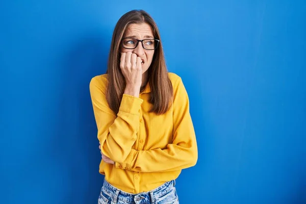 青い背景の上に立って眼鏡をかけている若い女性は 口の中に爪をかむ上で手で強調し 緊張している 不安の問題 — ストック写真