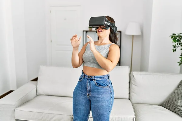 年轻美丽的惊慌失措的女人在家里用虚拟现实眼镜玩电子游戏 — 图库照片