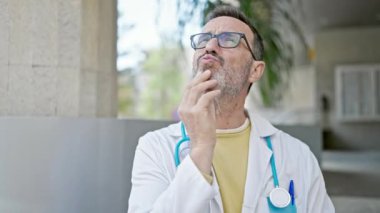 Orta yaşlı doktor şüpheli bir ifadeyle ayakta duruyor ve hastanede düşünüyor.
