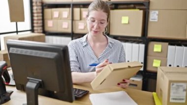 Genç sarışın kadın ecommerce iş işçisi. Ofiste paket üzerinde bilgisayar kullanıyor.