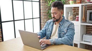 Sofrada dizüstü bilgisayar kullanan genç latin adam yemek odasında kazanan yüz ifadesiyle oturuyor.