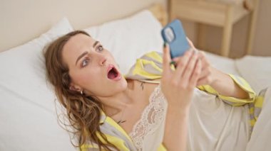 Yatak odasında uzanmış akıllı telefon kullanan genç sarışın kadın.