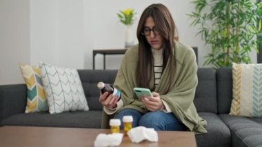 Genç, güzel, İspanyol bir kadın elinde ilaç şişesiyle evde akıllı telefon kullanıyor.
