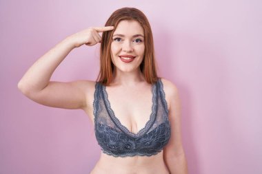 Pembe arka planda iç çamaşırı giyen kızıl saçlı kadın gülümseyerek tek parmağıyla kafasını işaret ediyor, harika bir fikir ya da düşünce, iyi bir hafıza. 