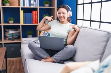 Evde dizüstü bilgisayar kullanan, kulaklık takan, kendine güvenen, gülümseyen, kendini gururlu ve mutlu gösteren İspanyol genç bir kadın.. 