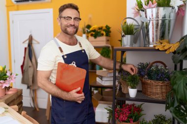 Middle age man florist touching plant holding clipboard at flower shop