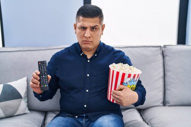 Latin asıllı genç adam TV 'de patlamış mısır yiyor şüpheci ve gergin, sorun yüzünden surat asıyor. negatif kişi. 