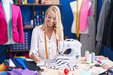 Young blonde woman tailor talking on smartphone writing on notebook at sewing studio