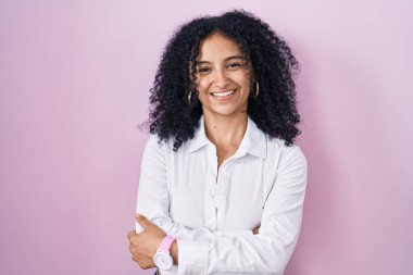 Hispanic woman with curly hair standing over pink background happy face smiling with crossed arms looking at the camera. positive person. 