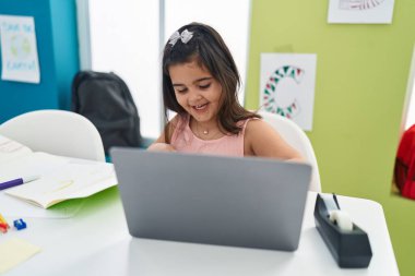 Adorable hispanic girl student using laptop sitting on table at classroom