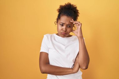 Sarı arka planda duran kıvırcık saçlı genç İspanyol kadın. Alnındaki bir sorun yüzünden endişeli ve stresli. Kriz için endişeli. 