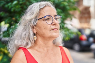 Orta yaşlı, kır saçlı bir kadın parkta ciddi bir ifadeyle yan tarafa bakıyor.