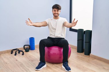 Sakallı İspanyol bir adam yoga odasında pilatese oturmuş kucaklaşmak için kollarını açmış kameraya bakıyor. Mutluluğu kucaklayan neşeli bir ifade.. 