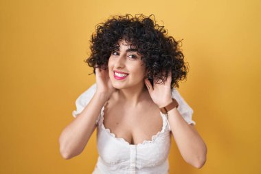 Sarı arka planda duran kıvırcık saçlı genç esmer kadın iki elini de kulak hareketi yaparken duymaya çalışıyor, dedikoduya meraklı. İşitme sorunu, sağır 