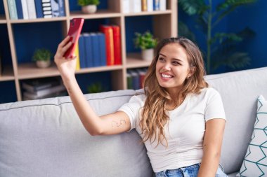 Young beautiful hispanic woman make selfie by smartphone sitting on sofa at home