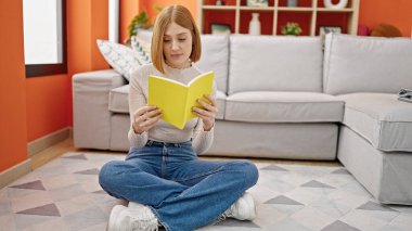 Kitap okuyan genç sarışın kadın evde yerde oturuyor.