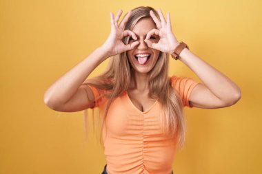Sarı arka planda duran genç bir kadın dürbün gibi jest yapıyor, dili dışarı çıkarıyor, gözleri parmaklarından bakıyor. çılgın ifade. 
