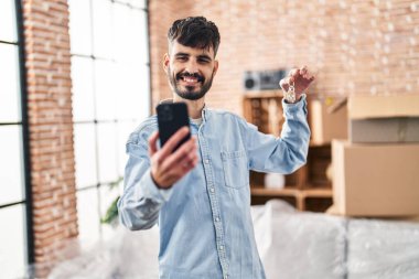 Genç İspanyol adam yeni evinde elinde anahtarla akıllı telefondan selfie çekiyor.