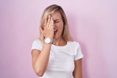 Young blonde woman standing over pink background yawning tired covering half face, eye and mouth with hand. face hurts in pain. 