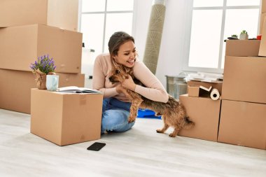 Young beautiful hispanic woman hugging dog sitting on floor at new home