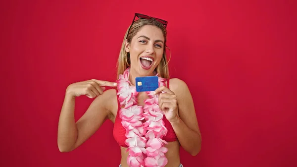 Young blonde woman tourist pointing to credit card smiling over isolated red background