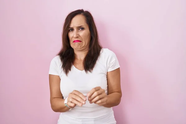 Middle age brunette woman standing over pink background disgusted expression, displeased and fearful doing disgust face because aversion reaction.