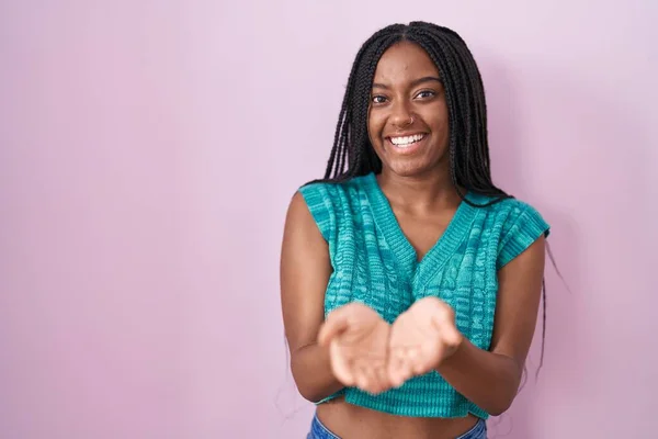 年轻的非洲裔美国人 头戴辫子 站在粉色背景上微笑着 双手合十 接受或给予手势 持有和保护 — 图库照片