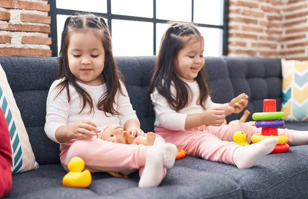 可爱的孪生姐妹们抱着娃娃坐在家里的沙发上玩圈圈游戏 — 图库照片