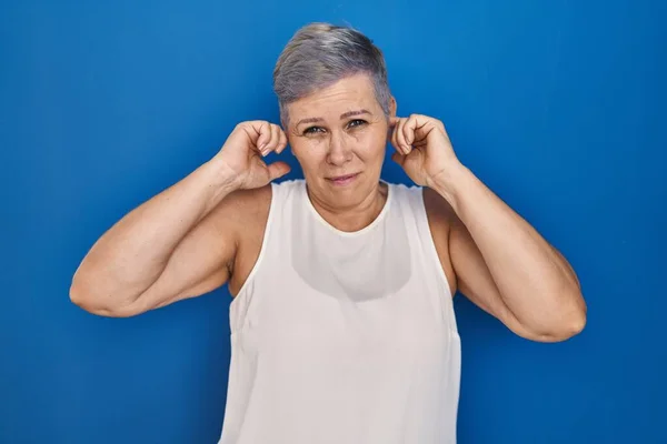 中年白人妇女站在蓝色背景之上 用手指捂住耳朵 对嘈杂的音乐感到恼怒 聋人概念 — 图库照片