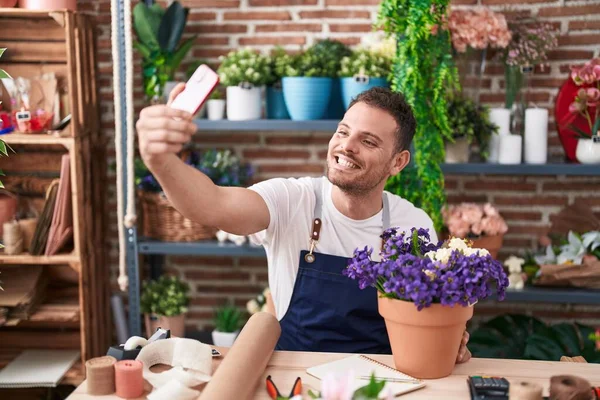 Young hispanic man florist make selfie by smartphone sitting on table at florist