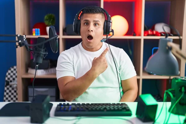 Молодой Латиноамериканец Играющий Видеоигры Удивлен Показывая Пальцем Сторону Открытый Рот — стоковое фото
