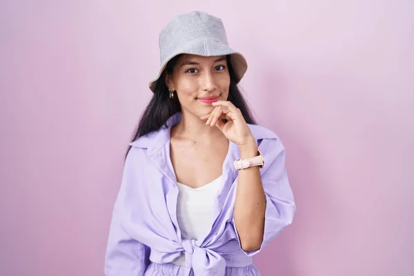 若いヒスパニック系の女性ピンクの背景の質問については 包括的な表現を考えて手で帽子をかぶって立っている 笑顔で思いやりのある顔 疑わしい概念 — ストック写真