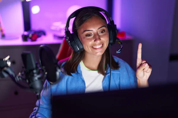 stock image Young hispanic woman wearing headphones playing video games smiling with an idea or question pointing finger with happy face, number one 