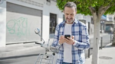 Orta yaşlı bir adam caddede motosiklet başında akıllı telefon kullanıyor.