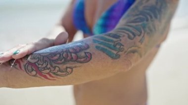 Orta yaşlı, kır saçlı, bikini giyen bir turist kumsalda koluna güneş kremi sürüyor.
