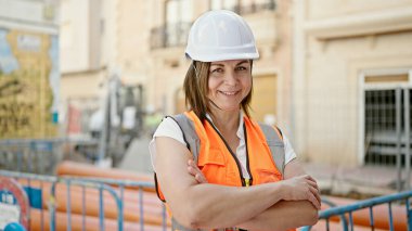 Orta yaşlı İspanyol kadın müteahhit inşaat alanında kollarını kavuşturup gülümsüyor.