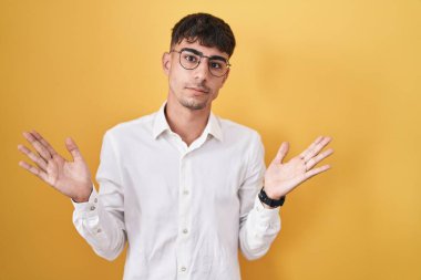 Young hispanic man standing over yellow background clueless and confused expression with arms and hands raised. doubt concept. 
