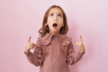 Pembe arka planda duran küçük İspanyol kız şaşırmış ve şaşırmış bir şekilde yukarı bakıyor ve parmaklarıyla işaret ediyor ve kollarını kaldırıyor.. 