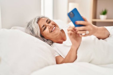 Orta yaşlı bir kadın yatakta akıllı telefon kullanıyor.