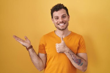 Young hispanic man standing over yellow background showing palm hand and doing ok gesture with thumbs up, smiling happy and cheerful 